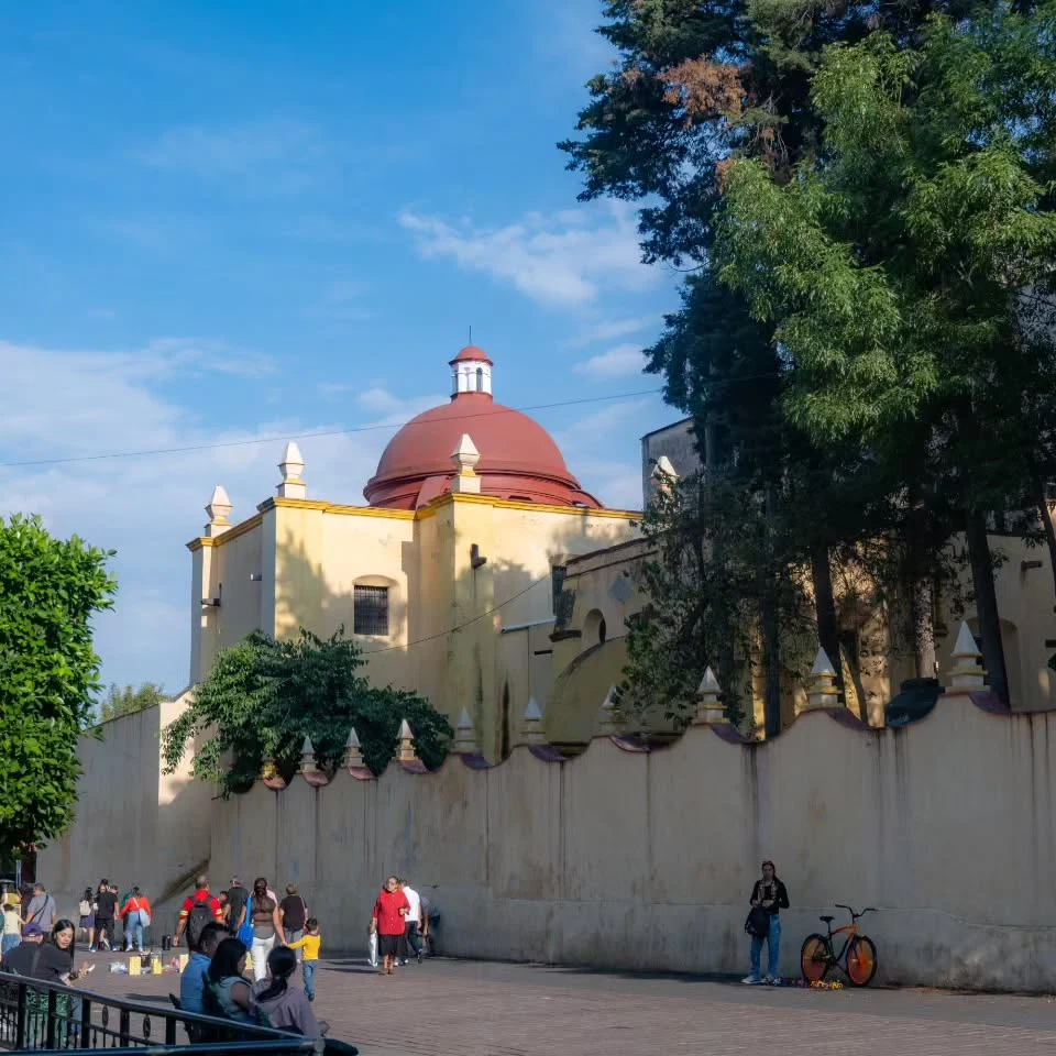 People beside a church with a red dome, beneath trees and a blue sky in Mexico.