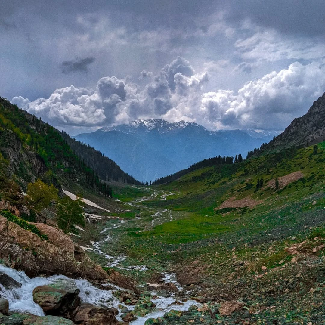 A rocky stream running through a green mountain valley beneath clouds.